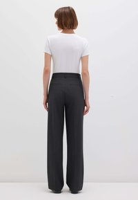 Donna con capelli corti e castani, che indossa una camicia bianca a maniche corte e pantaloni larghi grigi scuri a vita alta, in piedi di fronte a una parete bianca.