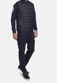 Navy puffer vest with horizontal stitching, front zipper, and logo on chest, paired with navy cargo pants and dark sneakers with a light sole.