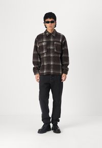 Chemise en flanelle à carreaux marron avec une texture subtile, deux poches sur la poitrine, jean noir et bottes noires à lacets. Des lunettes de soleil complètent le look.