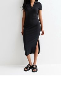 Schwarzes, geripptes Midikleid mit Kragen und kurzen Ärmeln, das einen Oberschenkel-hohen Schlitz aufweist. Kombiniert mit schwarzen, gestrapselten Sandalen.