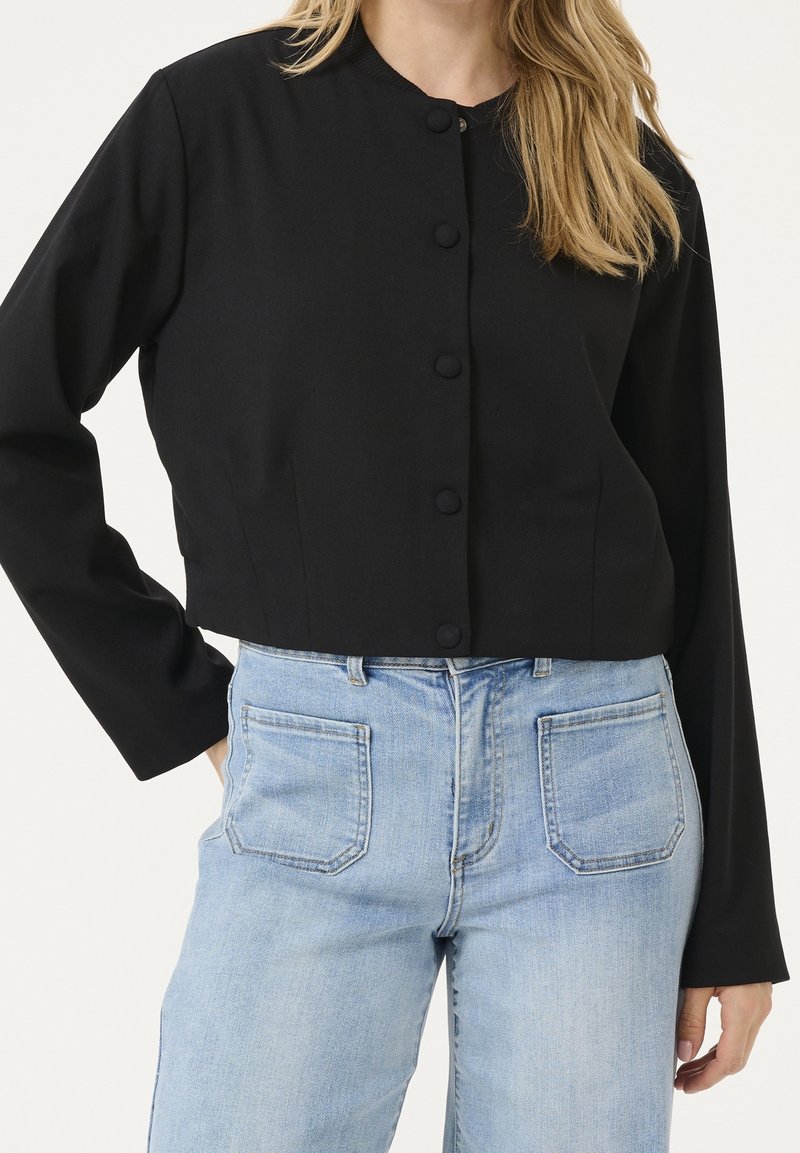 Femme portant une veste noire courte boutonnée à manches longues et un jean bleu clair taille haute avec poches avant visibles.
