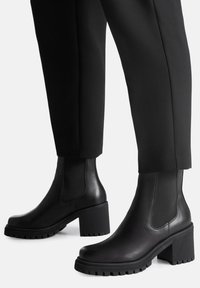 Czarne botki na obcasie z gładką skórzaną powierzchnią, elastycznymi panelami po bokach oraz grubymi, teksturowanymi gumowymi podeszwami zapewniającymi przyczepność i stabilność.