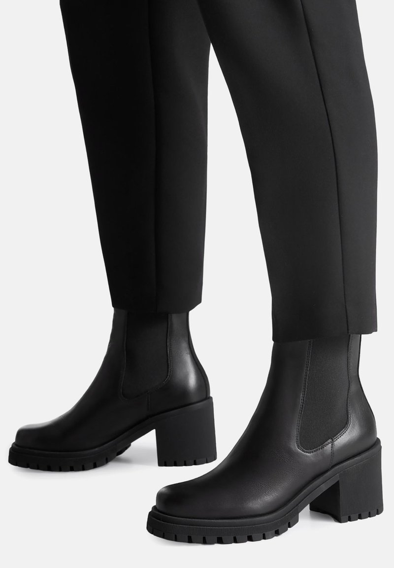 Czarne botki na obcasie z gładką skórzaną powierzchnią, elastycznymi panelami po bokach oraz grubymi, teksturowanymi gumowymi podeszwami zapewniającymi przyczepność i stabilność.