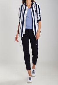 Randig bomberjacka i marinblått och vitt, lager på lager över en ljusblå topp. Svarta avsmalnande byxor och vita sneakers fulländar outfiten.