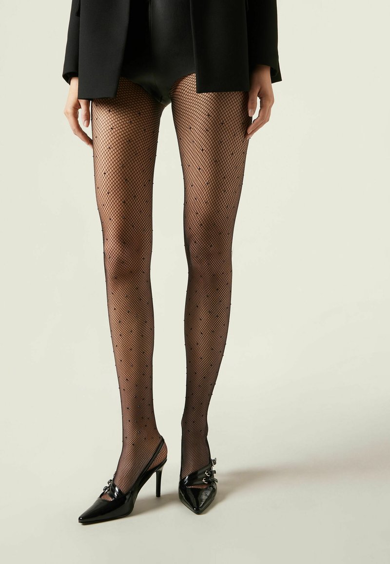 Tights de red negras con pequeños puntos negros, combinadas con tacones altos negros de punta con herrajes decorativos.