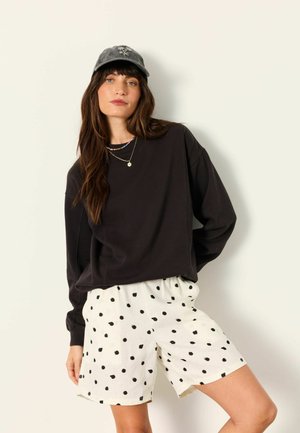 Woman wearing a black long-sleeve sweatshirt, white shorts with black polka dots, a gray cap, and layered necklaces, standing against a white wall.