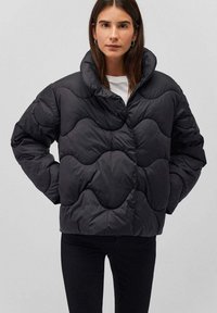 Giacca a vento nera con un design a righe trapuntato, colletto alto e chiusura a scatto, abbinata a una maglietta bianca e jeans neri.