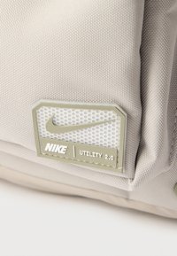 Κοντινό πλάνο ετικέτας υφάσματος Nike Utility 2.0 σε μπεζ χρώμα με το λογότυπο Nike και υφή φόντου πάνω σε υφή μπεζ υφάσματος.