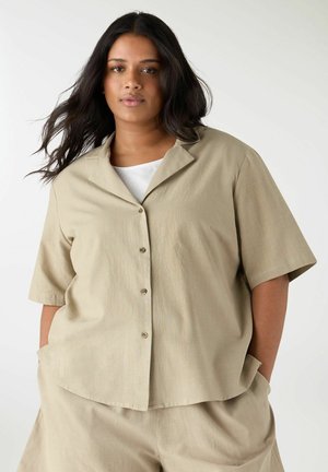 Donna taglia forte con capelli lunghi e scuri che indossa una camicia beige a maniche corte abbottonata e pantaloni abbinati, in piedi con le mani nelle tasche.