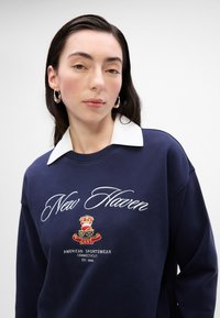 Navy bluza z białym kołnierzykiem, haftowanym napisem "New Haven" oraz detalem herbu w kolorach czerwonym i żółtym. Miękka tekstura materiału.