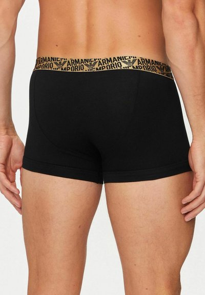 Uomo che indossa boxer aderenti neri con elastico in vita dorato con logo Emporio Armani ripetuto, mostrato di spalle.