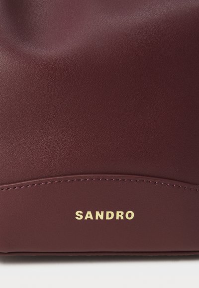 Borsa in pelle bordeaux con texture liscia e leggera luminosità. Presenta la scritta dorata "SANDRO" e un dettaglio cucito lungo il fondo.