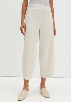 Donna con pantaloni crema a vita alta, a gamba larga e taglio corto con tasche laterali e scarpe crema abbinate senza lacci.