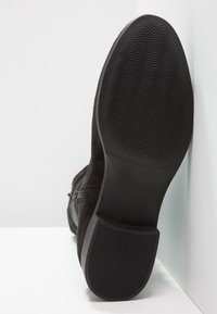 Suela de bota negra con patrones de relieve ondulados y lineales, tamaño 37 marcado cerca del centro, bota posicionada de pie sobre una superficie blanca.