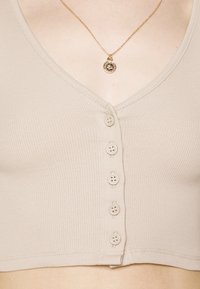 Crop top beige à côtes avec un décolleté en V et six boutons sur le devant ; comprend un délicat collier en or avec un pendentif circulaire.