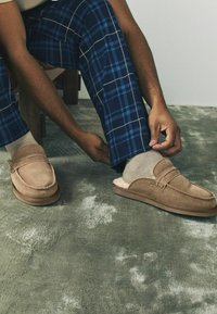 Osoba v modrých károvaných nohaviciach si upravuje papuče s béžovými ponožkami na štruktúrovanej šedej koberčekovej podlahe.