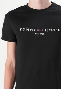 Camiseta de algodón negra con el texto blanco "TOMMY HILFIGER EST. 1985" y un acento rectangular rojo y azul. Diseño de mangas cortas y cuello redondo.