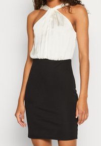 Robe style halter avec un haut plissé crème et une jupe noire ajustée. Texture lisse ; sans motifs visibles ni détails en métal.