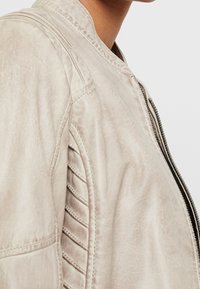 Světlá béžová bomber bunda s hladkým povrchem, žebrovanými bočními panely a kovovým zipem s malým táhlem.