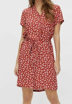 Mujer con vestido corto de manga corta, rojo con flores, con botones en la parte delantera y cinturón atado a la cintura, de pie frente a un fondo liso.