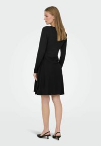 Femme en robe noire à manches longues, longueur genou, avec liens sur les côtés, et chaussures slingback noires à talons bas, debout de profil vers la gauche sur un fond uni.