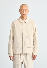Uomo con testa rasata che indossa una giacca e pantaloni beige abbinati, in piedi contro uno sfondo grigio chiaro uniforme.