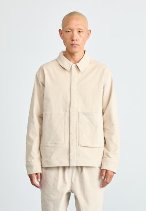 Man met een geschoren hoofd in een bijpassende beige knoopjasje en broek, staand tegen een plain lichtgrijze achtergrond.