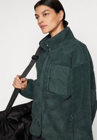 Grüne Fleecejacke mit strukturiertem Material, hohem Kragen und Reißverschlusstasche. Über einer schwarzen Tasche getragen, die glatte Stoffakzente aufweist.