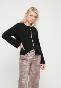 Black cardigan with white scalloped trim, long sleeves, and button closure. Paired with light pink pants featuring a tiger stripe pattern.