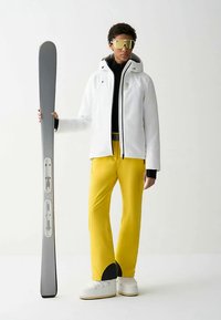Skidklädseln består av en vit isolerad jacka, svart underställ, gula byxor med svarta detaljer och en silverfärgad skida i handen. Guldtonade solglasögon fulländar looken.