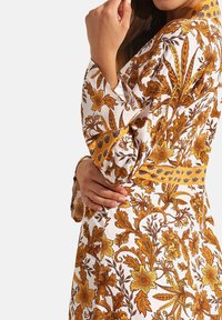 De bloemenprint robe heeft een witte stof met bruine en oranje botanische patronen, lange mouwen en een ceintuur in de taille, wat de textuur en het ontwerp accentueert.