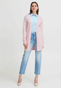 Cardigan rosa chiaro con apertura frontale, maniche lunghe e tasche, indossato sopra una camicia azzurro chiaro. Abbinato a jeans a vita alta e tacchi.