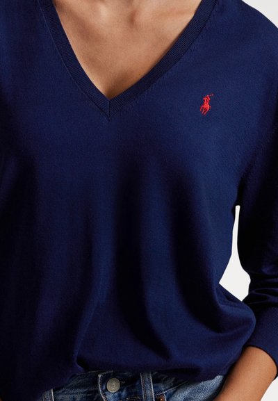 Camisola de gola em V azul-marinho, feita de tecido macio, com um logótipo vermelho bordado no peito esquerdo e mangas compridas. Usada com jeans de ganga.