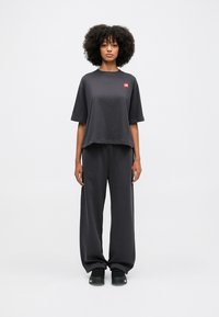 Crna pamučna majica kratkih rukava s crvenom logotip oznakom, u kombinaciji s širokim crnim hlačama i crnim tenisicama. Ukupno glatka tekstura.