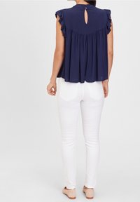 Donna in piedi, che indossa un top senza maniche blu navy con volant e schiena a oblò, pantaloni bianchi aderenti con tasche posteriori e scarpe beige.