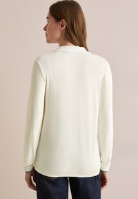 Blusa a maniche lunghe di colore crema con una texture liscia, dotata di collare e dettagli blu ai polsini, mostrata da un lato posteriore.