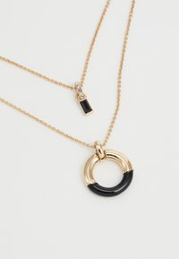 Colliers superposés en ton or avec un pendentif circulaire aux accents noirs et un charme rectangulaire orné de pierres claires.