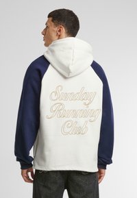 Sudadera con capucha en blanco y azul marino, con capucha, mangas raglán y "Sunday Running Club" bordado en dorado en la parte posterior.