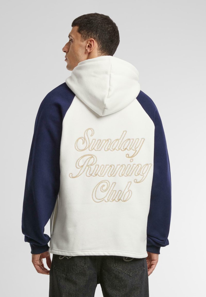 Sudadera con capucha en blanco y azul marino, con capucha, mangas raglán y "Sunday Running Club" bordado en dorado en la parte posterior.