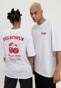 Deux personnes portant des t-shirts blancs surdimensionnés avec des graphismes de fruits rouges "Delicious", l'une vue de face et l'autre montrant le design au dos.