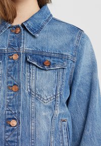 Džínová bunda světle modré barvy s klasickým límcem, měděnými knoflíky, předními kapsami a viditelnými švy na ramenou a švech.
