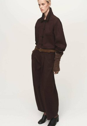 Camicia tailored marrone scuro con colletto, maniche oversize, due tasche sul petto, vita con cintura, pantaloni larghi abbinati e stivaletti neri con tacco.