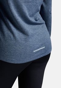 Maglietta sportiva leggera di colore blu realizzata in tessuto texturizzato, con una striscia riflettente sul bordo, abbinata a leggings neri.
