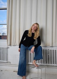 Cardigan nero con bottoni, jeans larghi in denim blu, a piedi nudi su uno sgabello metallico, con uno sfondo di tende neutre e una finestra.