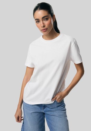 Kobieta o ciemnych włosach związanych w kucyk nosi jednolitą białą koszulkę i jasnoniebieskie dżinsy, stoi na szarym tle.