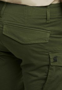 Pantaloni cargo verde oliva in tessuto, con multiple tasche, cuciture strutturate e una patch nera con logo sul lato.
