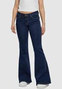 Uitlopende denim jeans in donkerblauw met gele stiksels, voorzien van een knoopsluiting, zijzakken en een licht versleten textuur aan de randen.