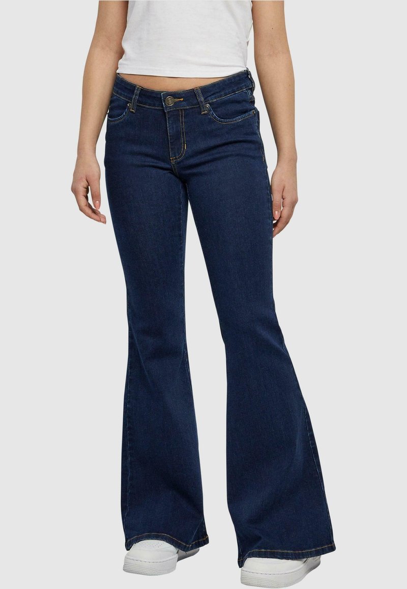 Uitlopende denim jeans in donkerblauw met gele stiksels, voorzien van een knoopsluiting, zijzakken en een licht versleten textuur aan de randen.