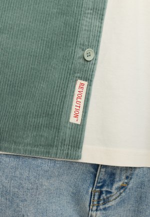 Groene corduroy kraag met knoop, wit katoenen shirt en spijkerbroek. Het label zegt "REVOLUTION" in het rood op de kraag.
