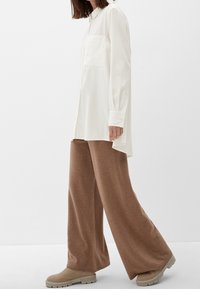 Biała koszula z długim rękawem, z kołnierzykiem i kieszonką, w połączeniu z szerokimi brązowymi dzianinowymi spodniami oraz jasnobeżowymi butami z teksturowaną podeszwą.